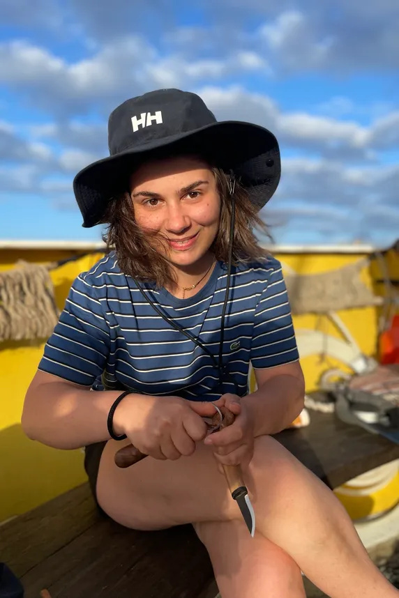 Alice Cherki sitting on deck in a striped shirt and sun hat, smiling, carving a wooden spoon with a knife.