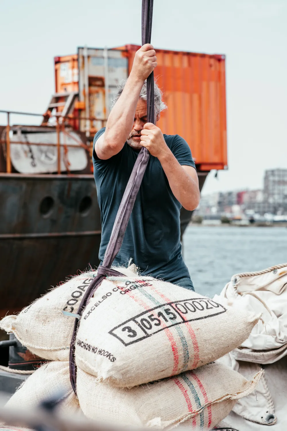 A crew member hoisting a burlap sack stamped 'Excelso Colombia Organic' dockside, orange shipping containers and a city skyline behind