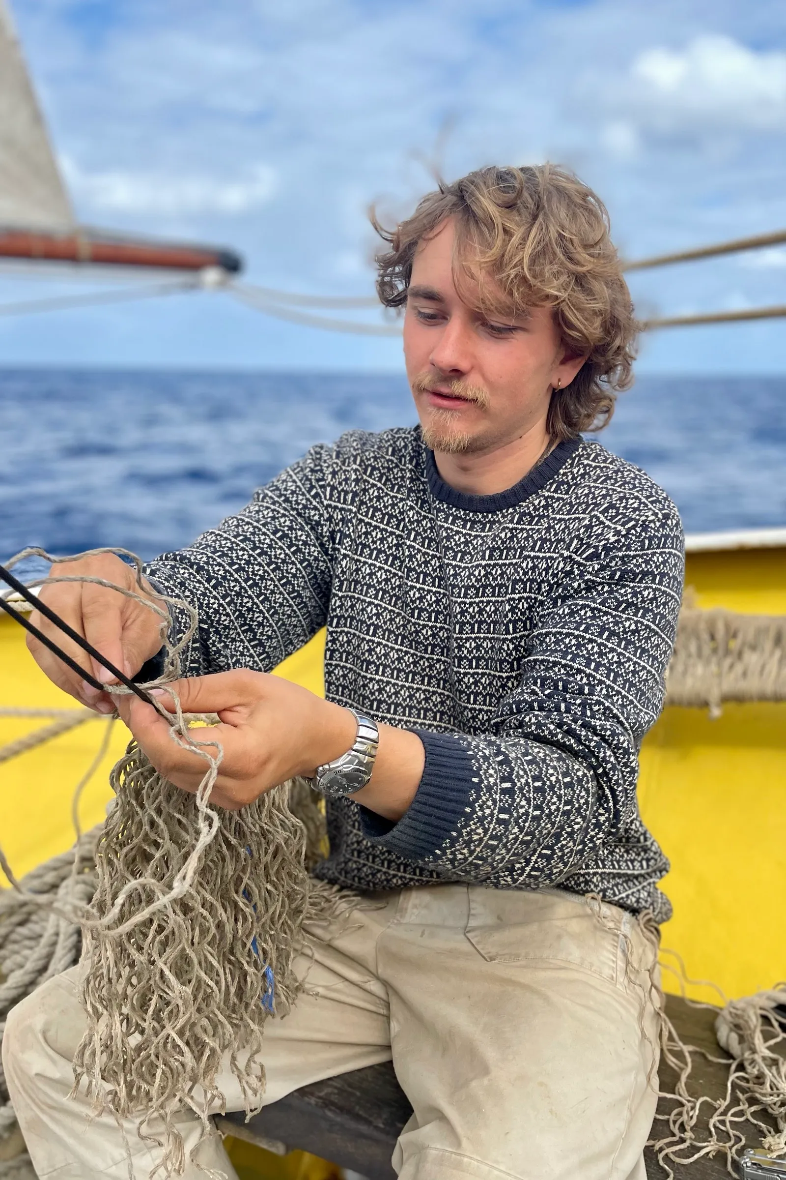 A young sailor sitting on deck in a wool sweater, splicing a rope net by hand with a fid, open ocean and sail behind him