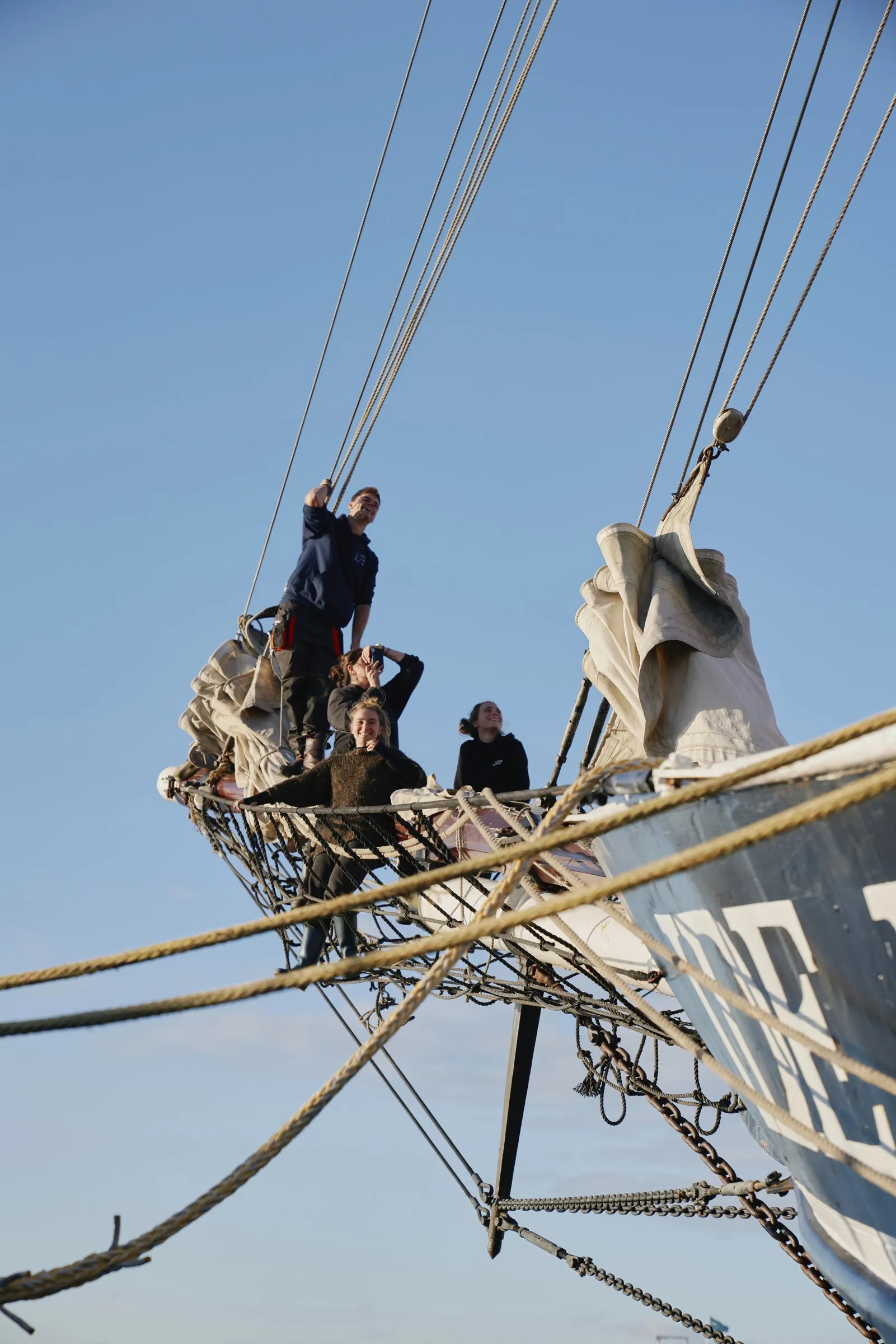 Four crew out on the bowsprit netting against a clear sky, one standing at the stay, the furled headsails stowed alongside them