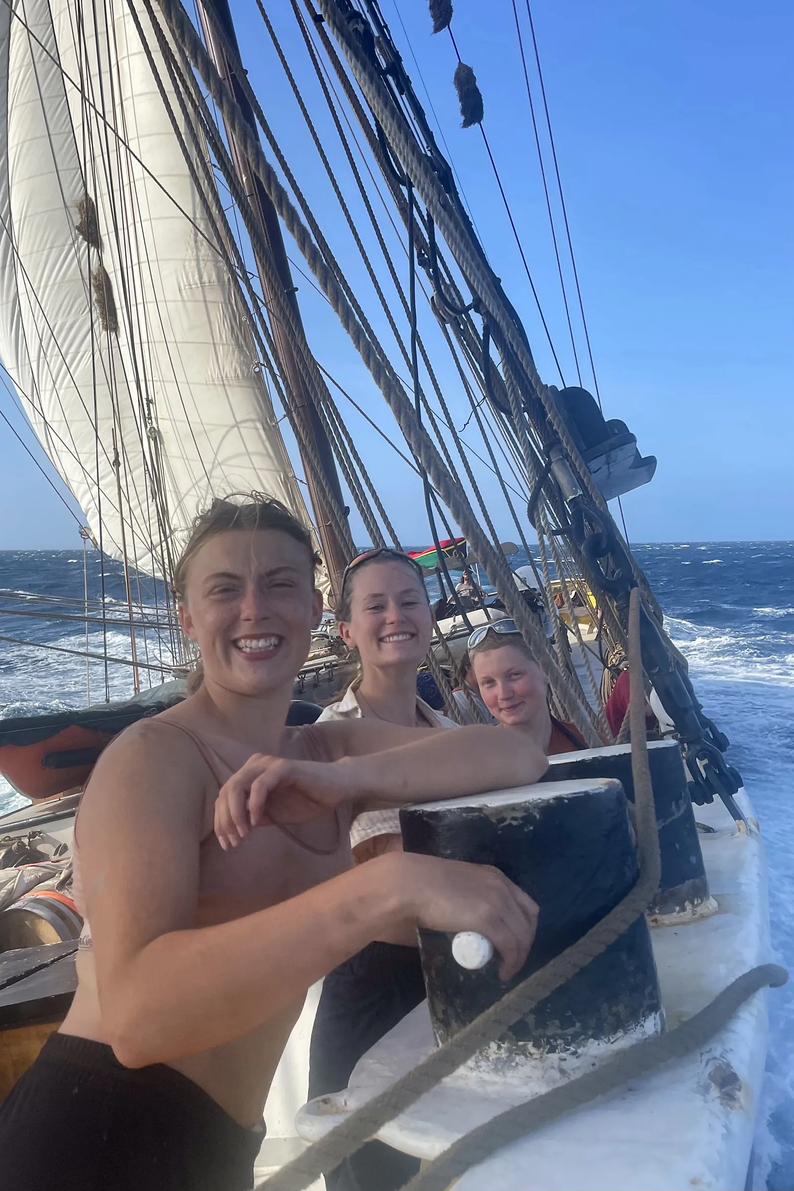 Three crew smiling by the rail as the ship heels through open blue water under full sail, the wake breaking alongside