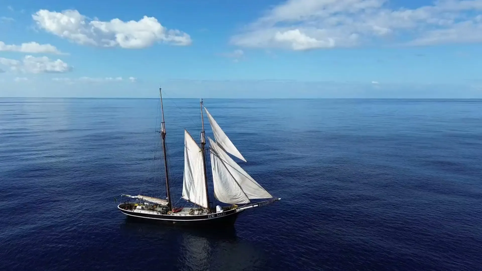 Aerial view of Ide Min under full sail on open water, her name legible on the hull