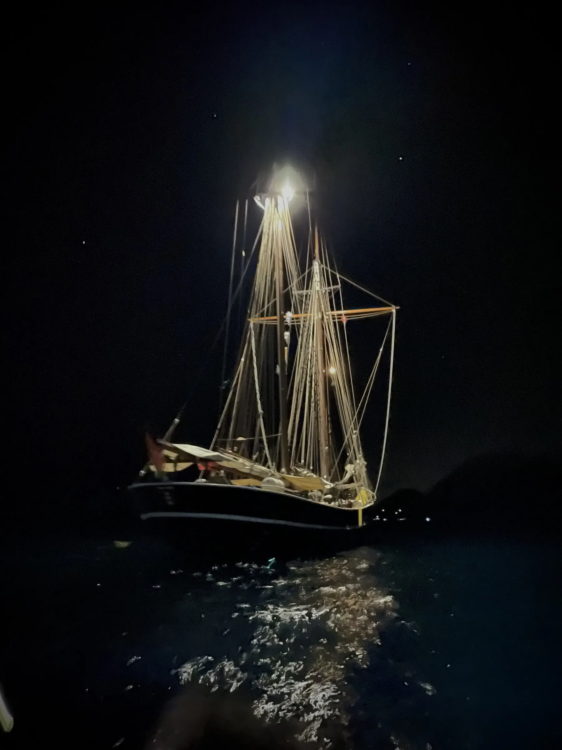 Ide Min at anchor after dark, her masthead light burning and the rigging lit warm against the black, the reflection breaking on the water