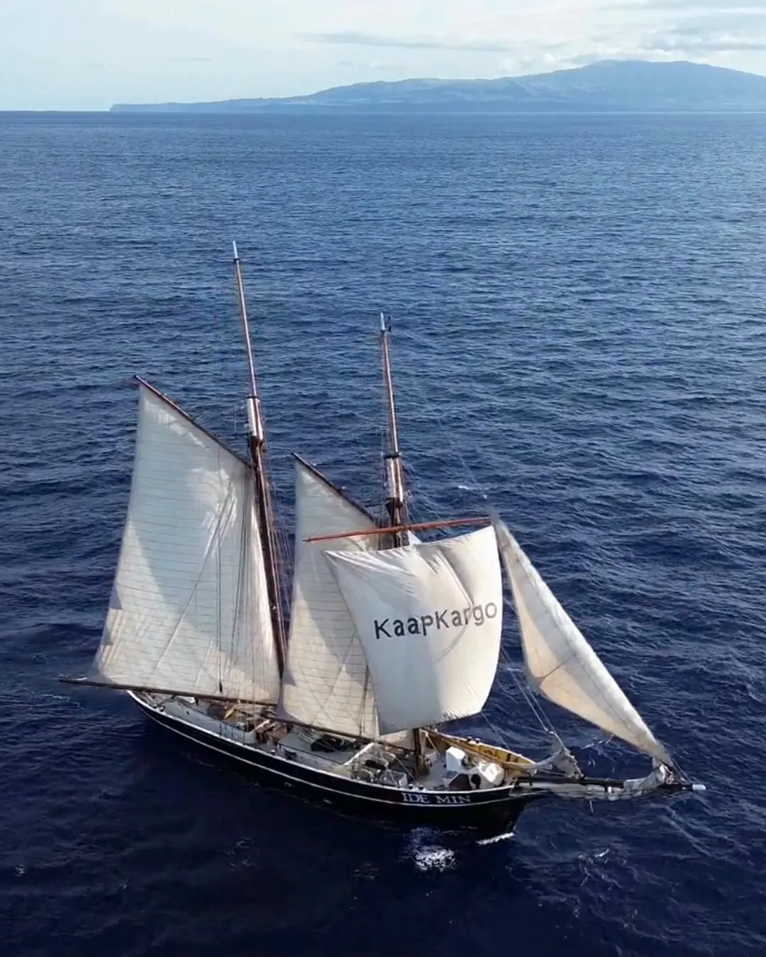 Ide Min under full sail seen from above, Kaap Kargo lettered on her foresail, a mountainous island on the horizon