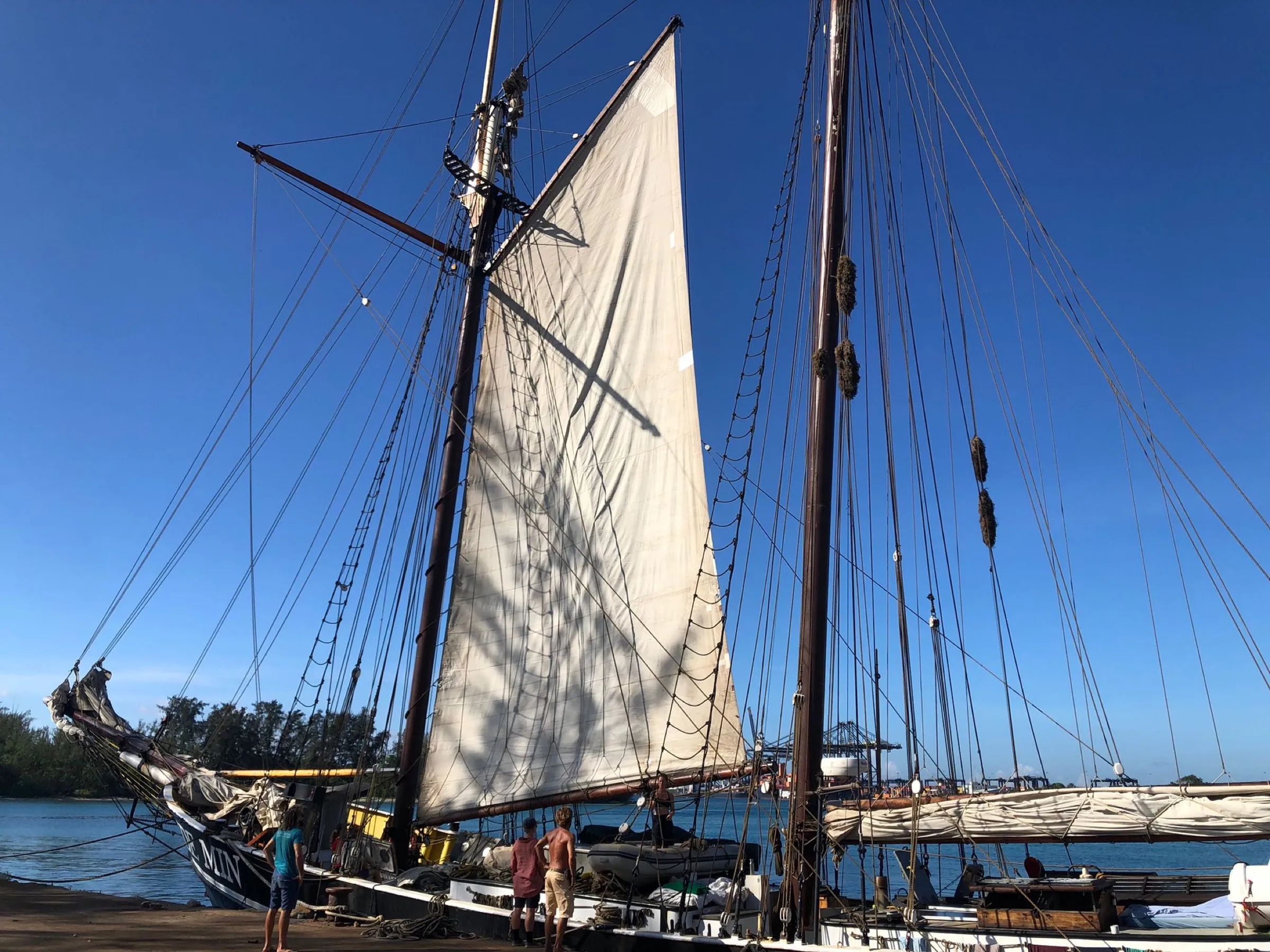 Ide Min moored at a quay under a blue sky, her gaff sail set to dry, three crew on the quay and deck, container cranes in the distance