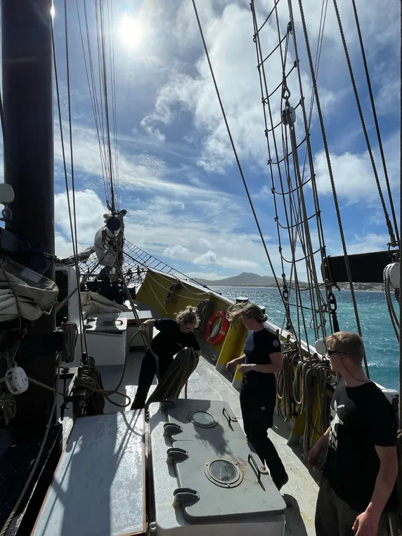 Crew coiling line on deck as Ide Min gets under way, Porto Santo's hills in the background.