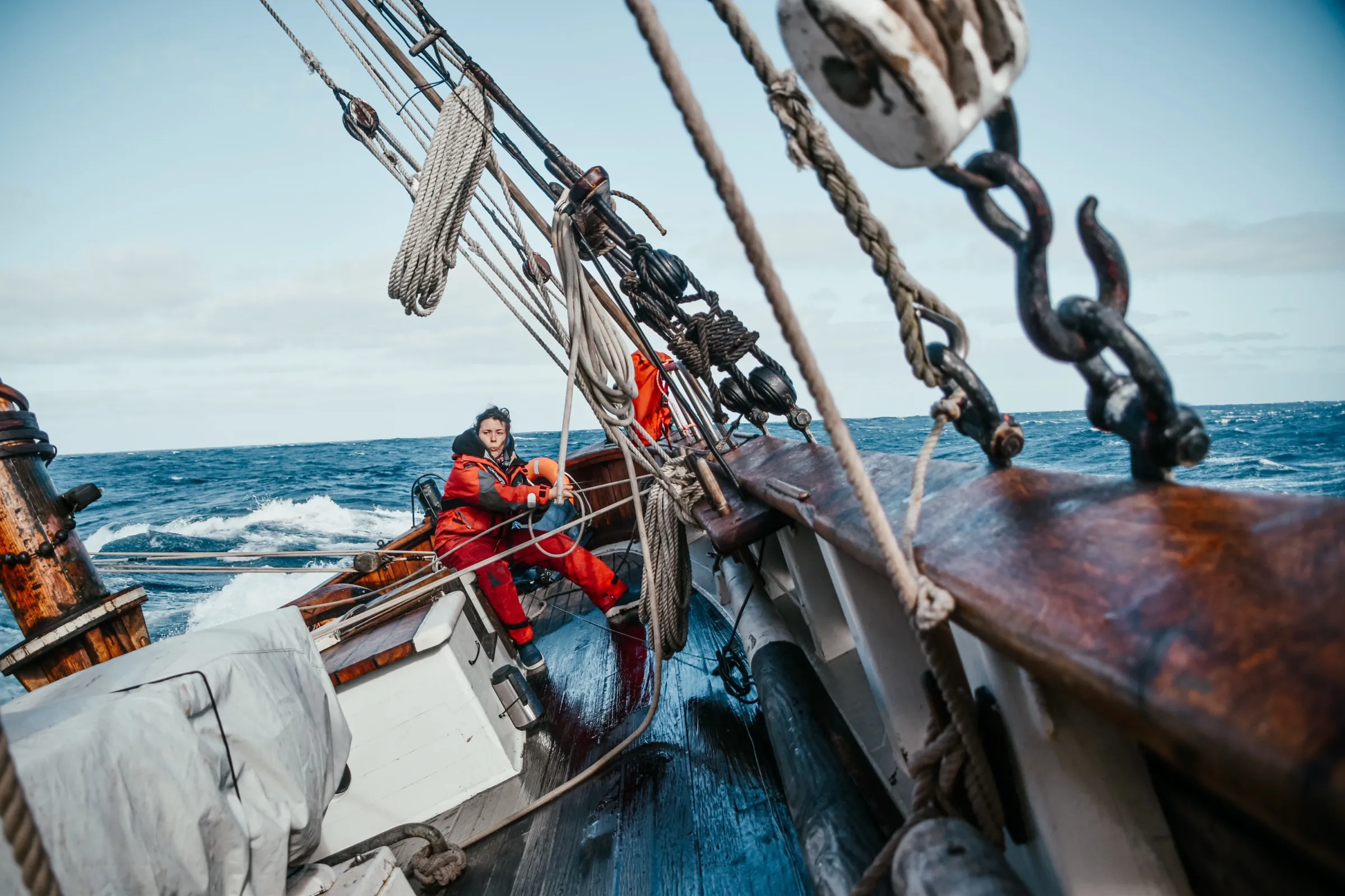 LUN II's crew in red foul-weather gear at the rail in a grey seaway, anchor gear and bowsprit in frame.