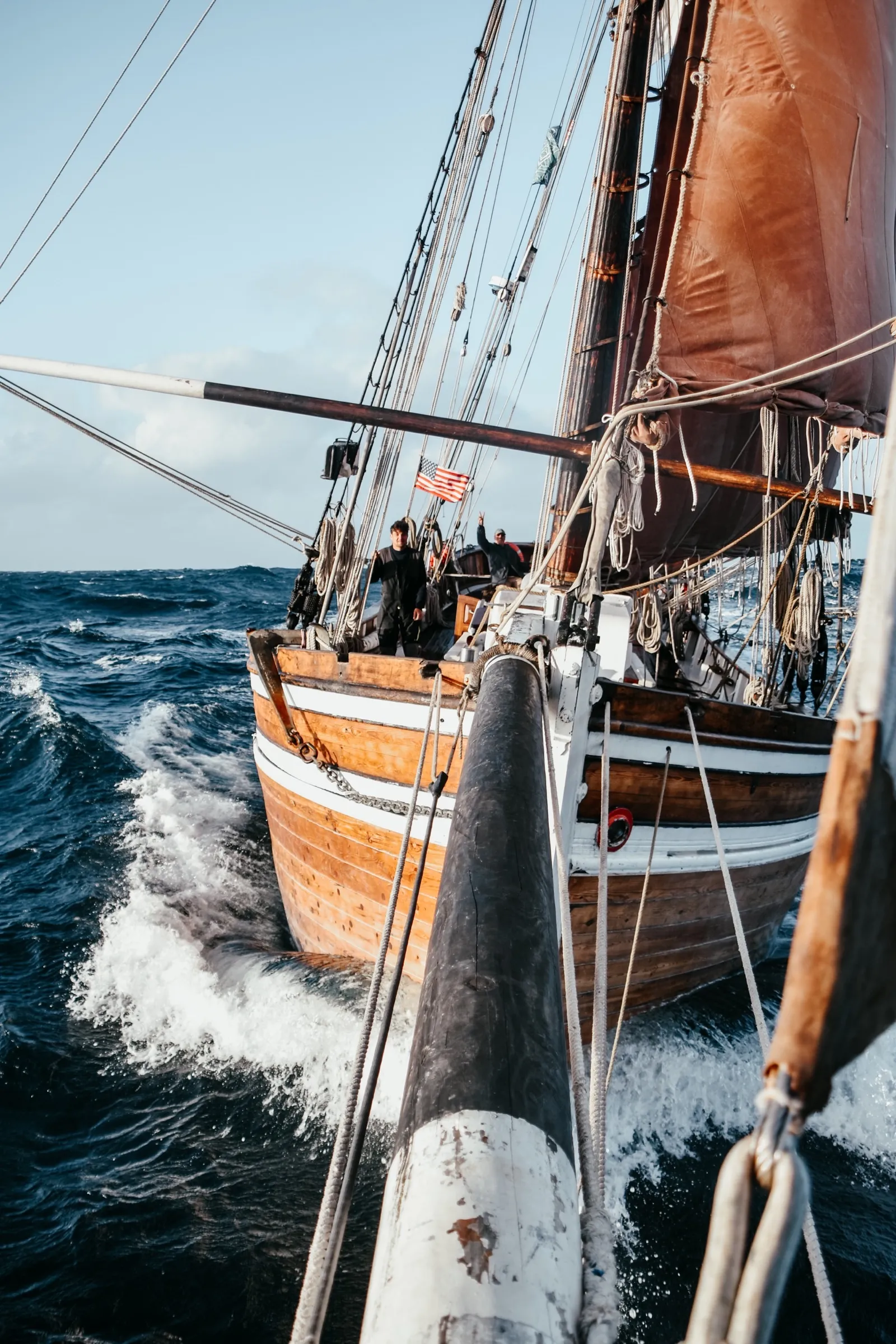 LUN II under sail on open water, seen from close alongside: her wooden hull and rigging, crew at work on deck, white water breaking away from her side