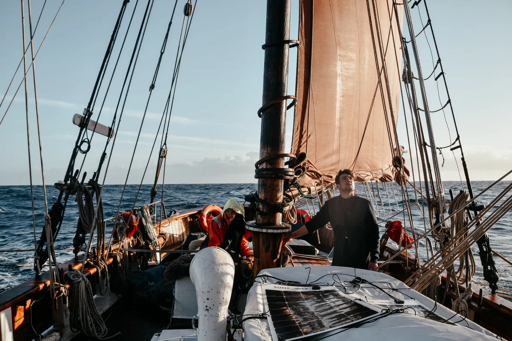 Golden-hour view from the foredeck of an orange headsail and solar panels, with two crew at work