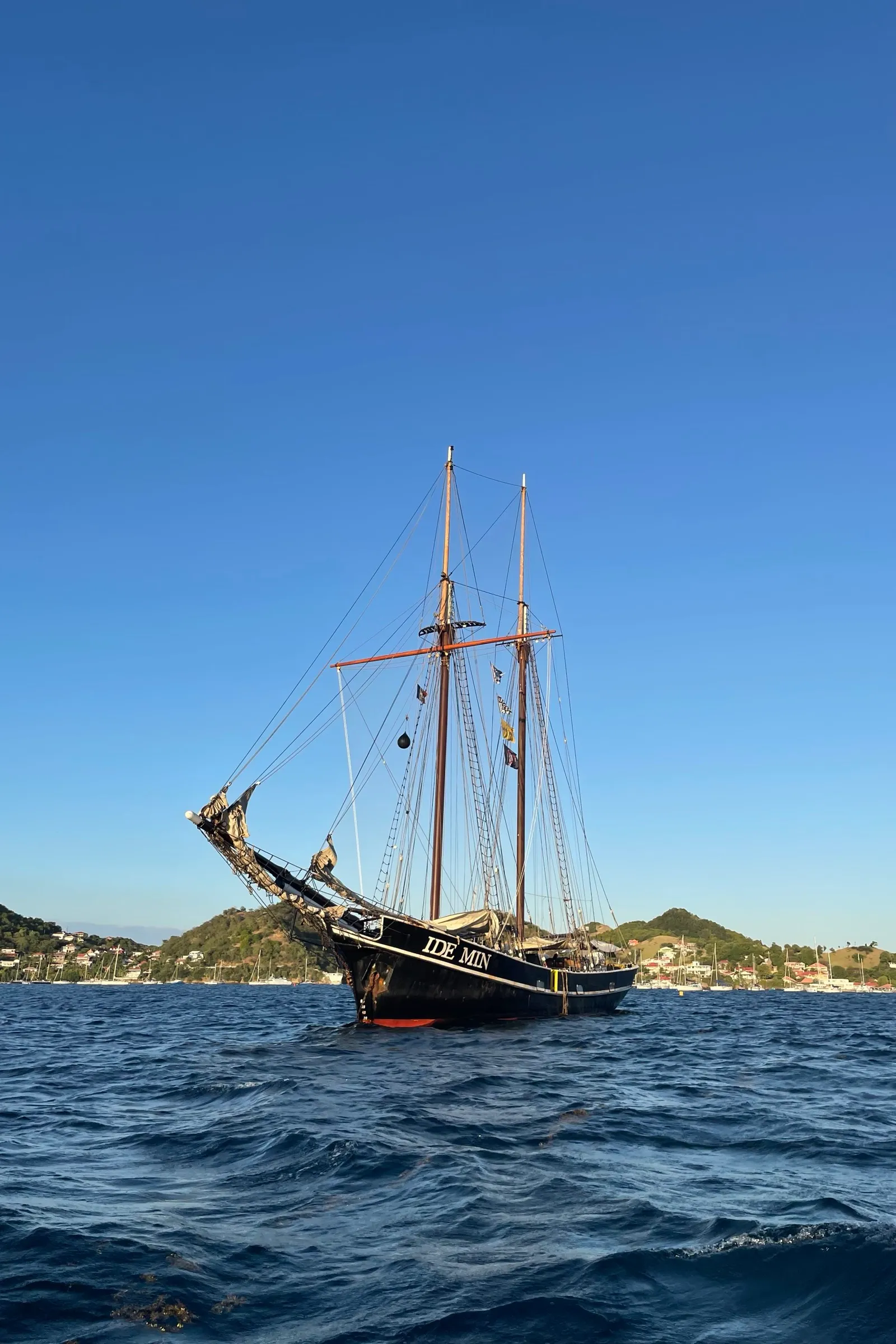 Ide Min lying at anchor in a Caribbean bay, her name on the bow, green hills and a small harbour full of yachts behind her